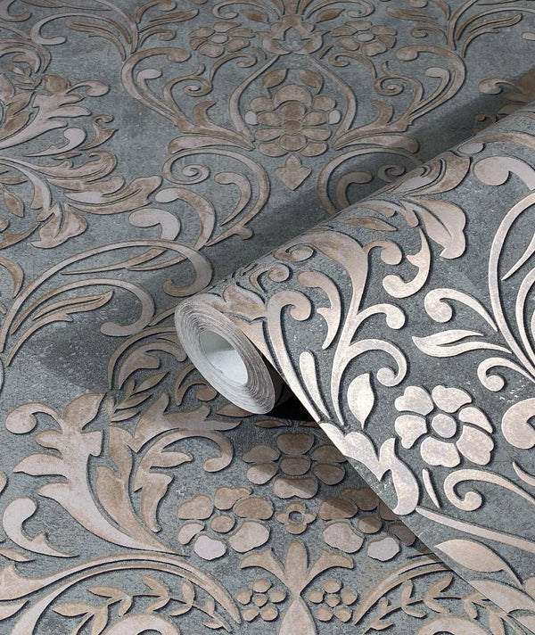 Marburg City Glam 32604 Close-up of a wallpaper roll on a matching surface, showing a large ornate damask floral and scroll pattern in light beige on gray.