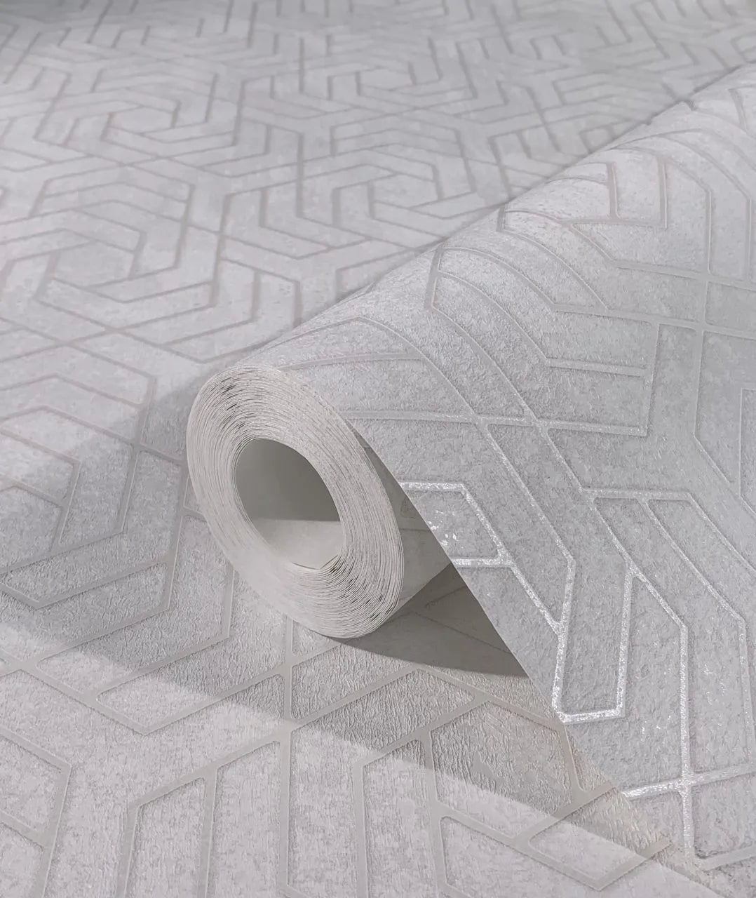 Marburg City Glam 32606 Close-up of a gray geometric patterned wallpaper roll resting on a matching unrolled sheet, showing embossed angular lines.