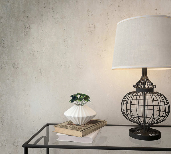 Marburg City Glam 32613 Light gray mottled wallpaper with a subtle plaster look behind a wire-frame table lamp, a small white planter with greenery, and books on a glass-topped table.