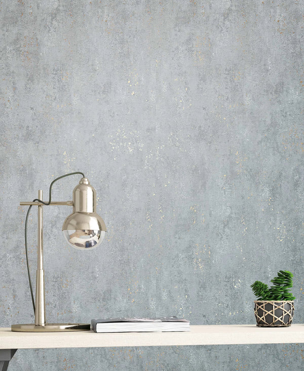 Marburg City Glam 32615 Gray textured wallpaper with faint metallic speckles shown behind a silver desk lamp, a small stack of magazines, and a potted plant on a light shelf.