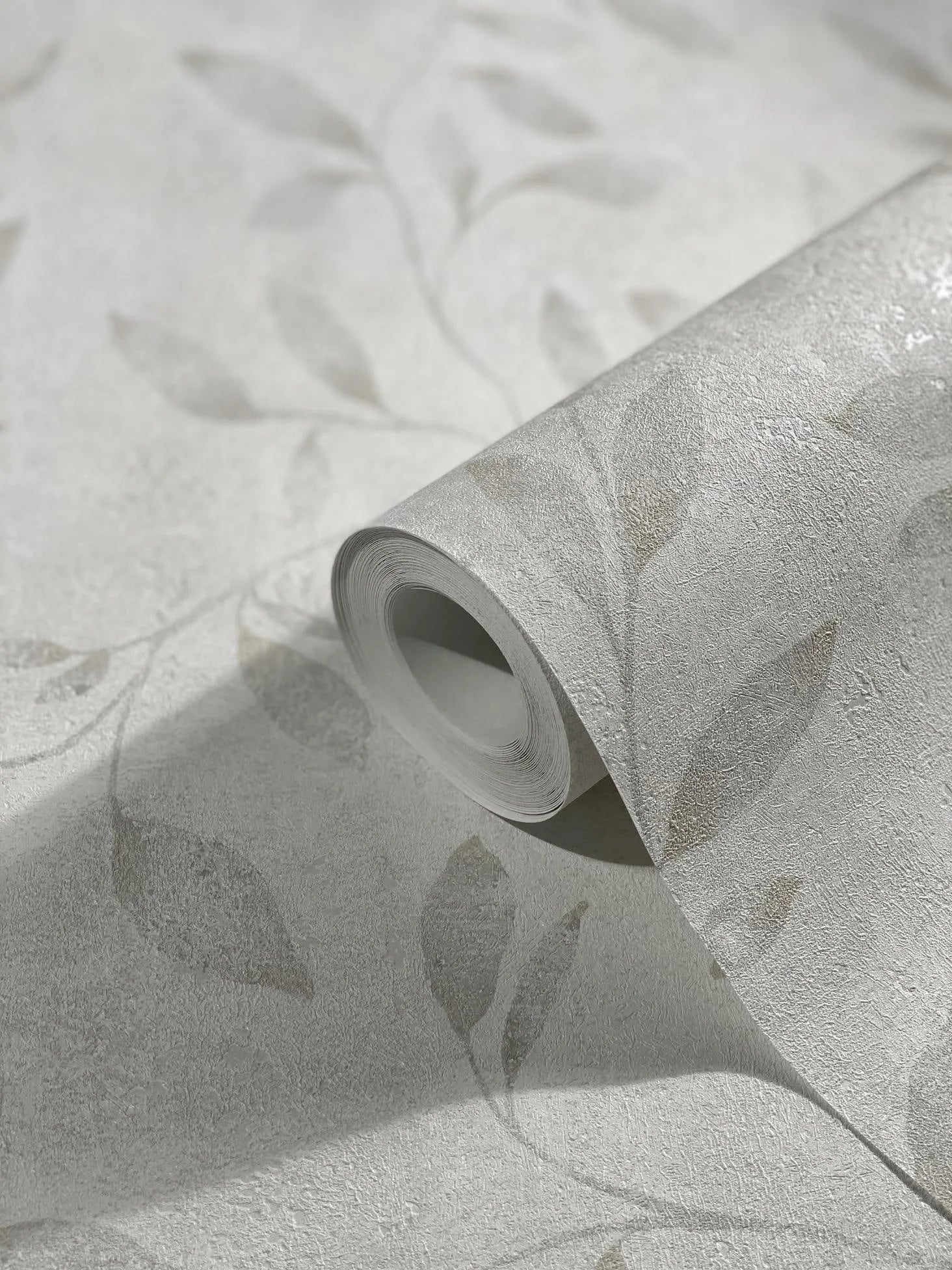 Marburg City Glam 32632 Close-up of a wallpaper roll resting on matching paper, showing a light neutral textured surface with a subtle tonal leaf pattern.