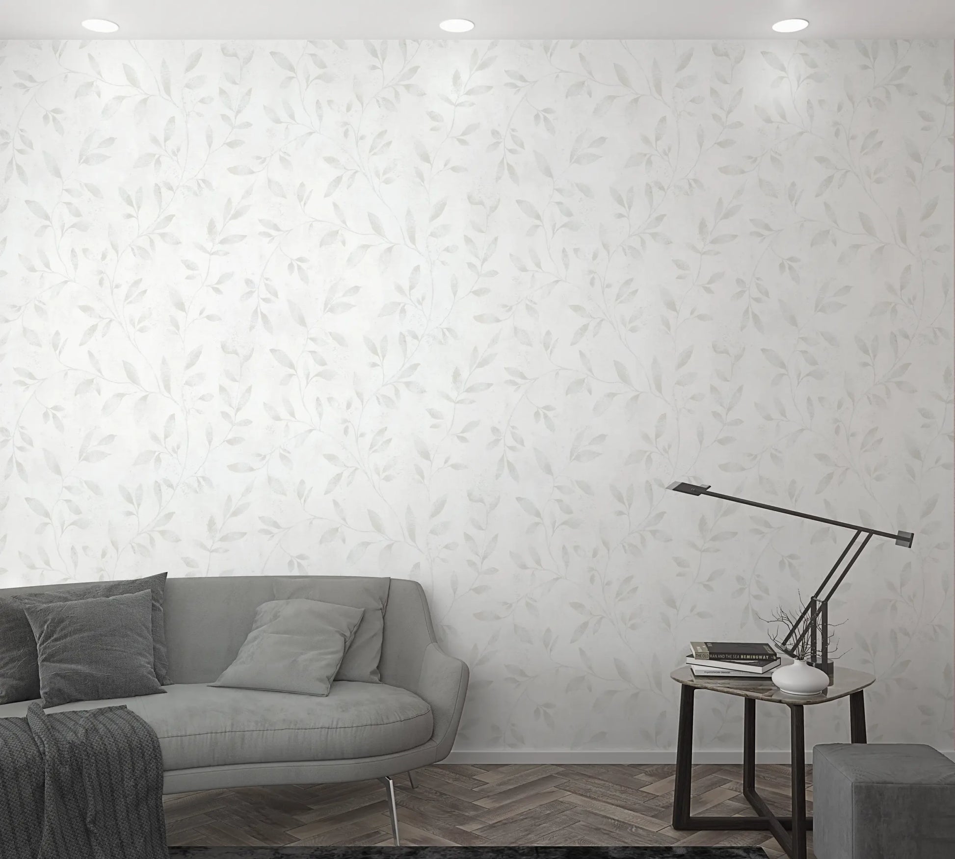 Marburg City Glam 32632 White wallpaper with a soft light-gray leafy vine pattern shown in a living room scene with a gray sofa, pillows, a side table with a slim desk lamp, books, and a small vase.