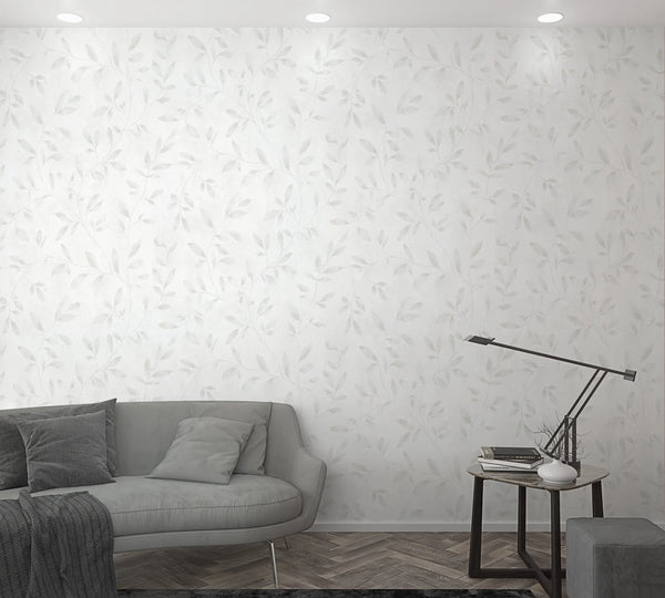 Marburg City Glam 32632 White wallpaper with a soft light-gray leafy vine pattern shown in a living room scene with a gray sofa, pillows, a side table with a slim desk lamp, books, and a small vase.