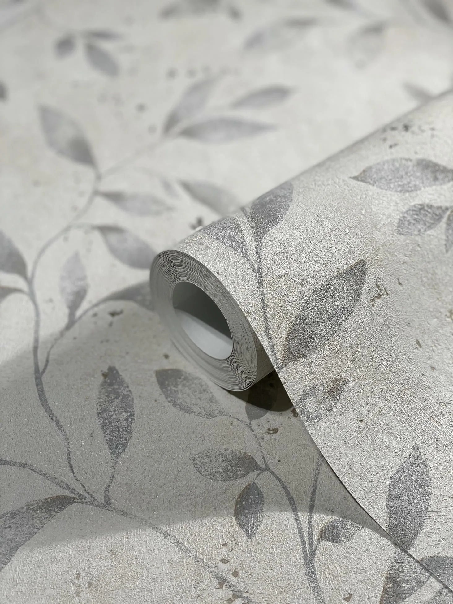 Marburg City Glam 32633 Close-up of a wallpaper roll resting on matching unrolled wallpaper, showing a textured light-neutral background with muted gray leafy vine pattern and slight speckling.