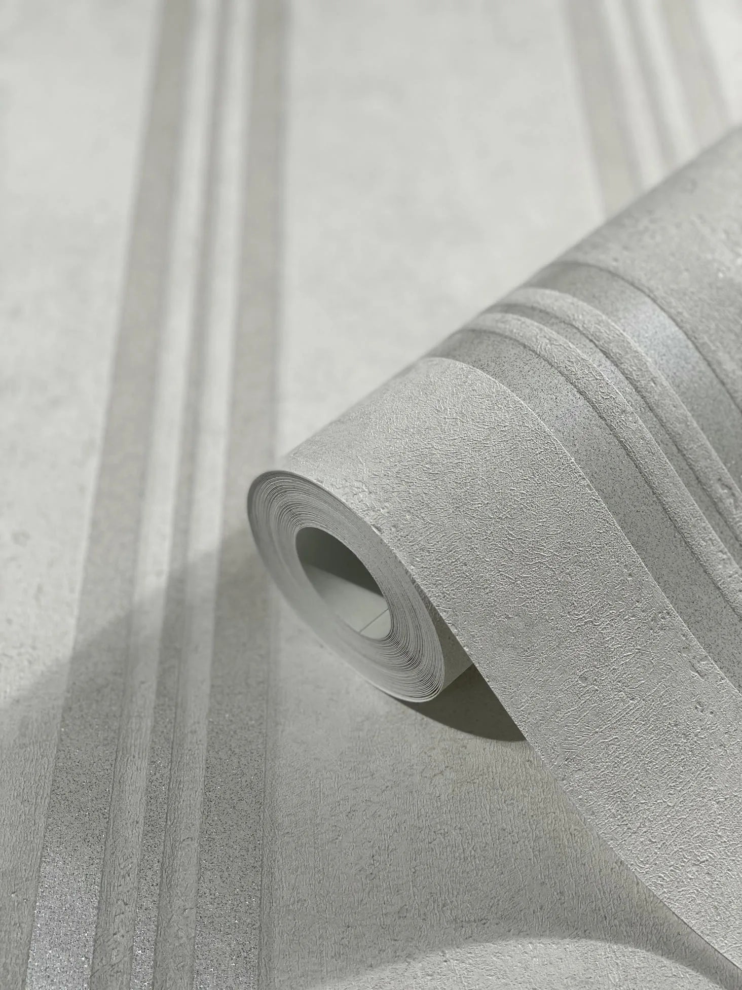 Marburg City Glam 32635 Close-up of a wallpaper roll on matching wallpaper showing a light gray textured surface with vertical tone-on-tone stripes and subtle shimmering bands.