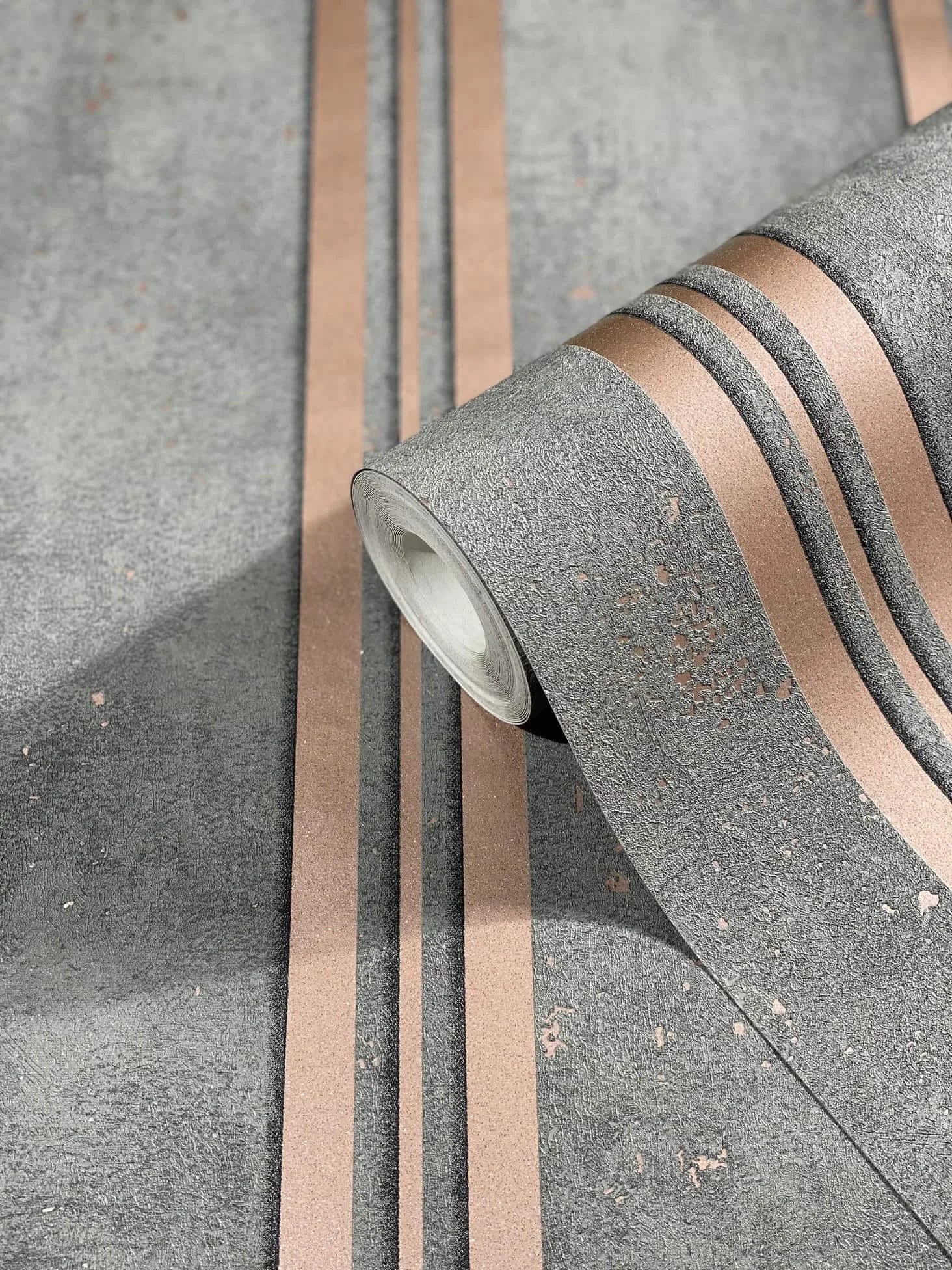 Marburg City Glam 32637 Close-up of a wallpaper roll on a matching surface, showing a textured grey ground with metallic copper parallel stripes and scattered distressed flecks.