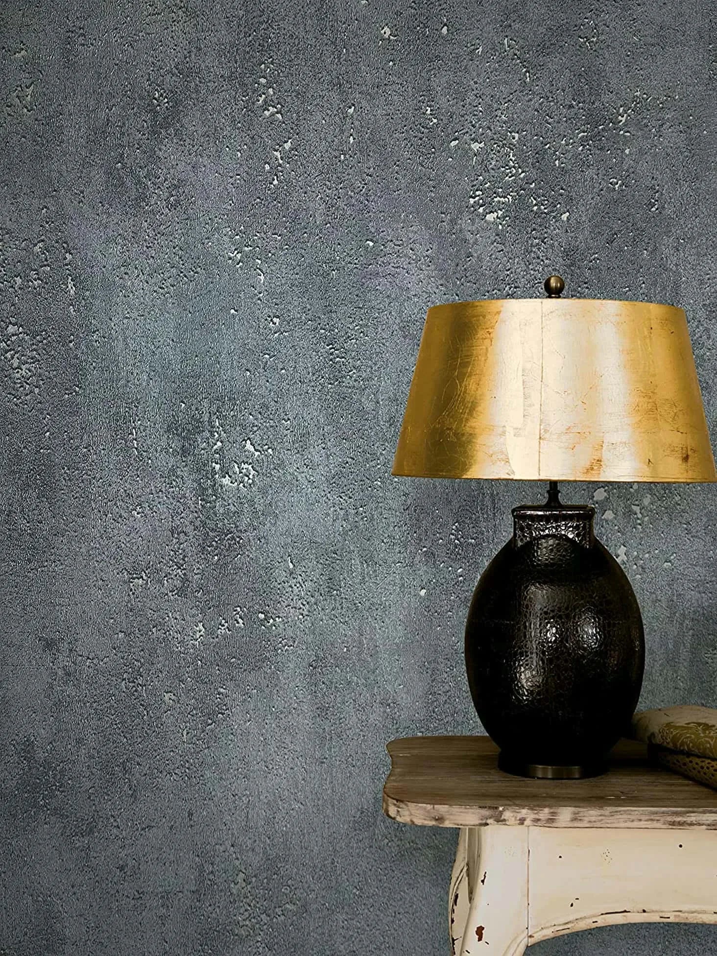 Marburg City Glam 32638 Textured dark grey concrete-look wallpaper behind a black table lamp with a gold shade on a worn wood side table.