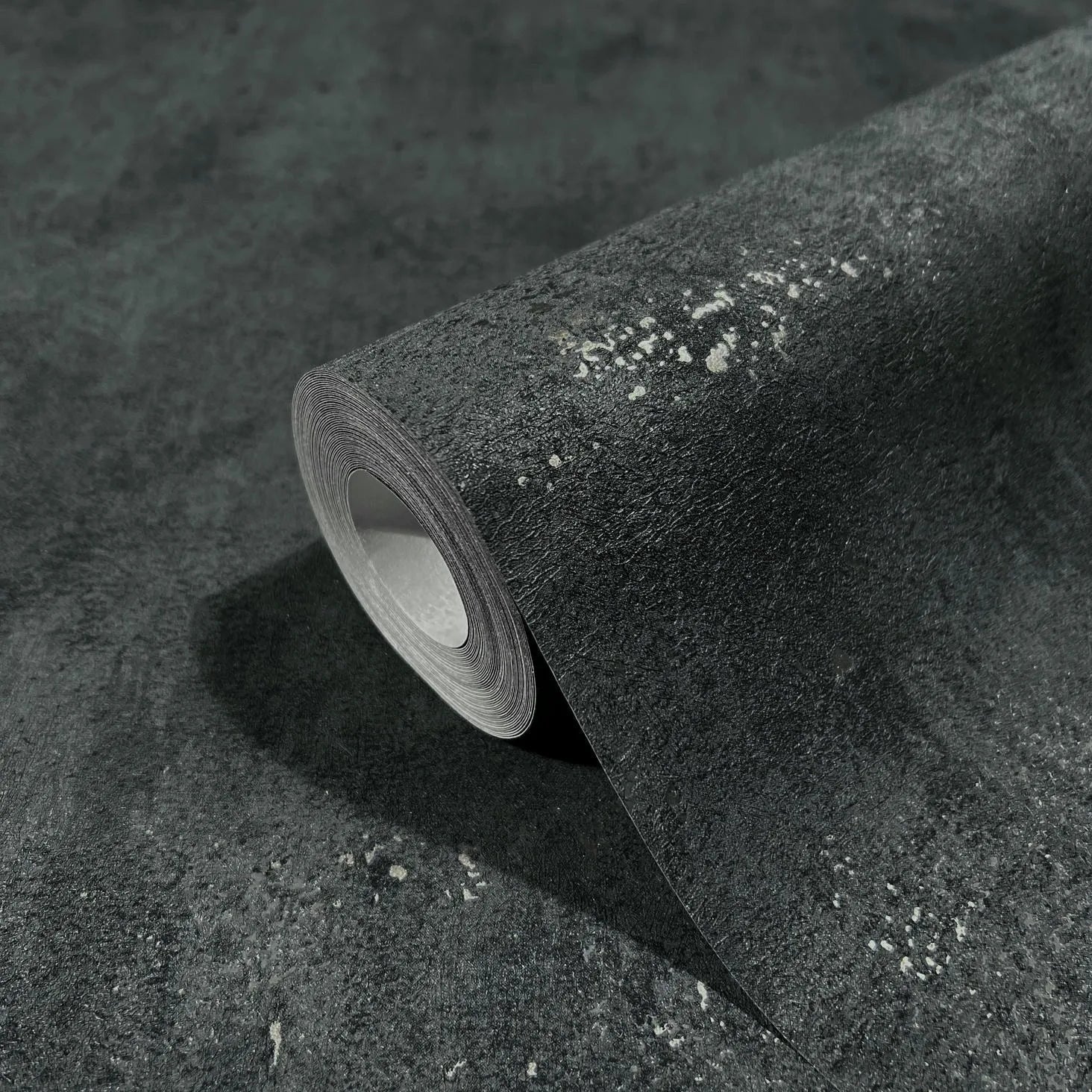 Marburg City Glam 32638 A dark mottled wallpaper roll with scattered light speckles, shown resting on a matching unrolled sheet.