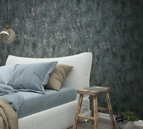 Marburg City Glam 32638 Dark charcoal textured wallpaper with scattered silver-like speckles behind a bed with blue bedding and beige pillows, a rustic wooden stool, a hanging glass pendant light, and a small potted plant.