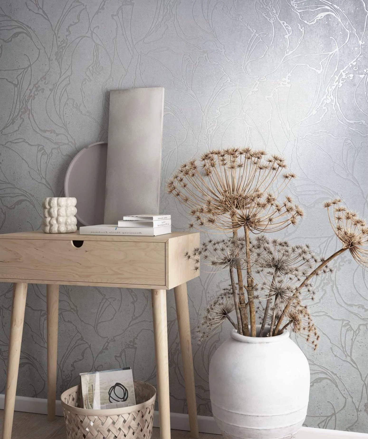 Marburg City Glow 34251 Light grey wallpaper with a large abstract botanical outline pattern shown behind a pale wood console table, a white ceramic vase holding dried stems, a woven basket, and small decor items.