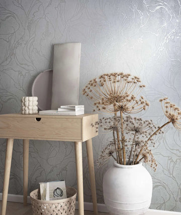 Marburg City Glow 34251 Light grey wallpaper with a large abstract botanical outline pattern shown behind a pale wood console table, a white ceramic vase holding dried stems, a woven basket, and small decor items.