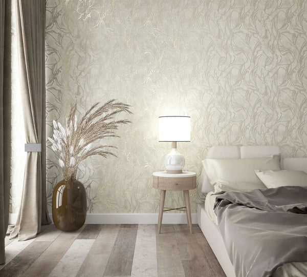 Marburg City Glow 34252 Bedroom scene showing light beige wallpaper with a flowing botanical pattern behind a bedside table with a white lamp, an upholstered bed with bedding, curtains, and a large vase holding pampas grass.
