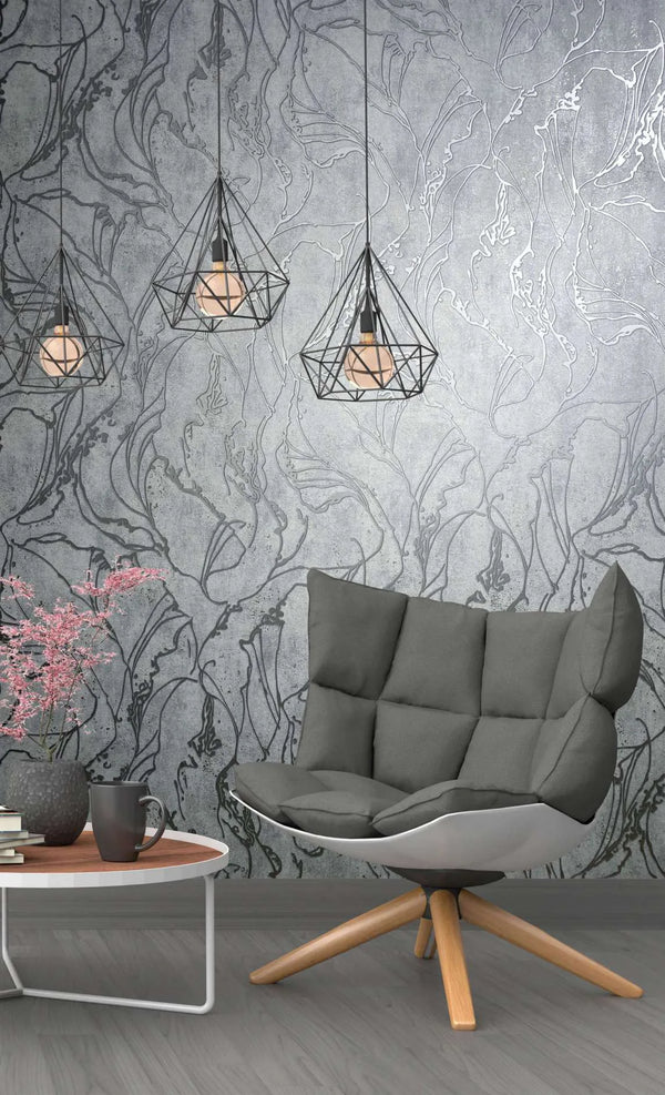 Marburg City Glow 34254 Gray wallpaper with large abstract botanical outlines on a mottled background, shown with geometric pendant lights, a cushioned lounge chair, and a small side table with books, a mug, and a vase of pink flowers.
