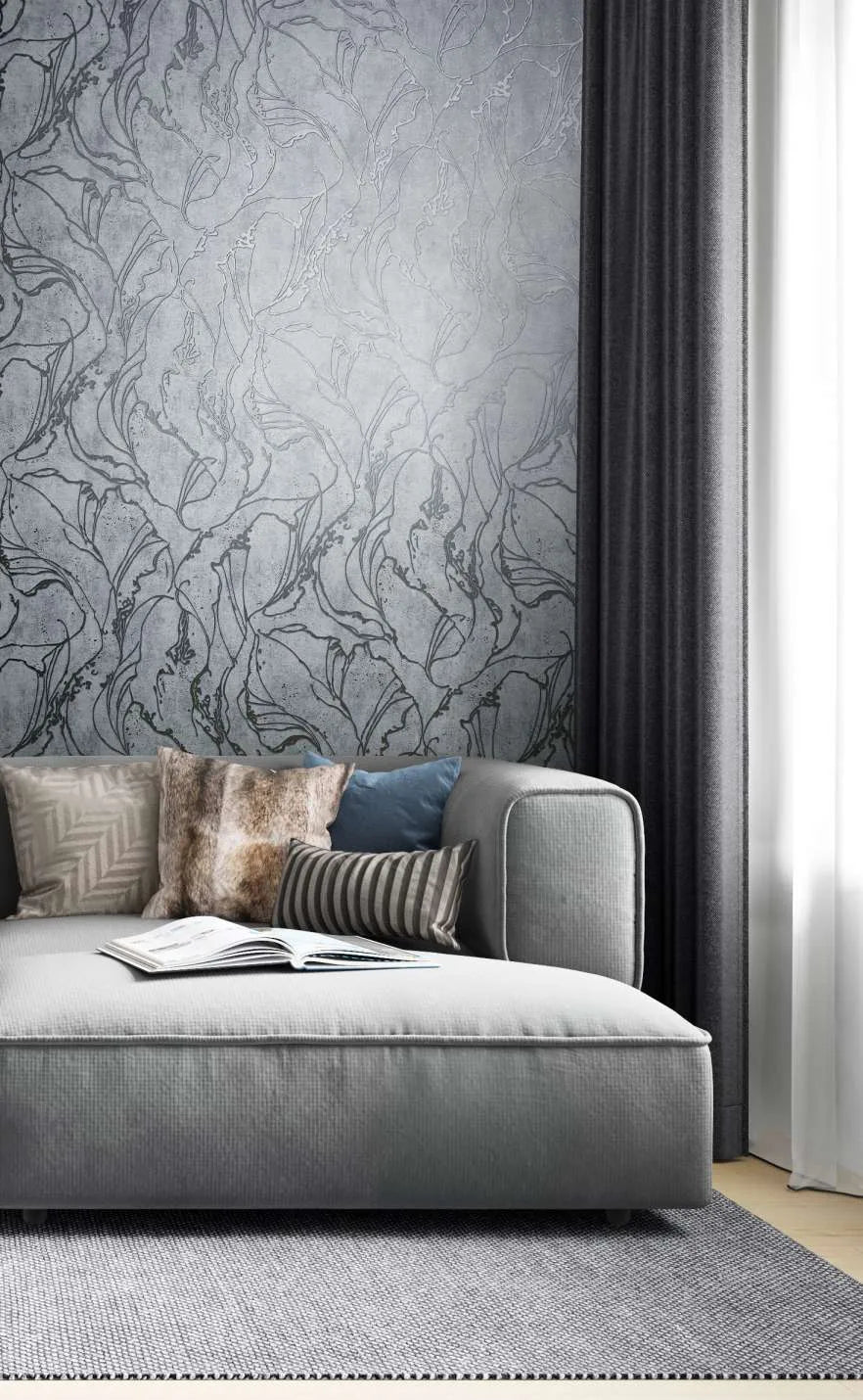 Marburg City Glow 34254 Gray wallpaper with a large flowing botanical line pattern behind a sofa with mixed pillows, next to dark curtains and a light rug; an open book rests on the seat.