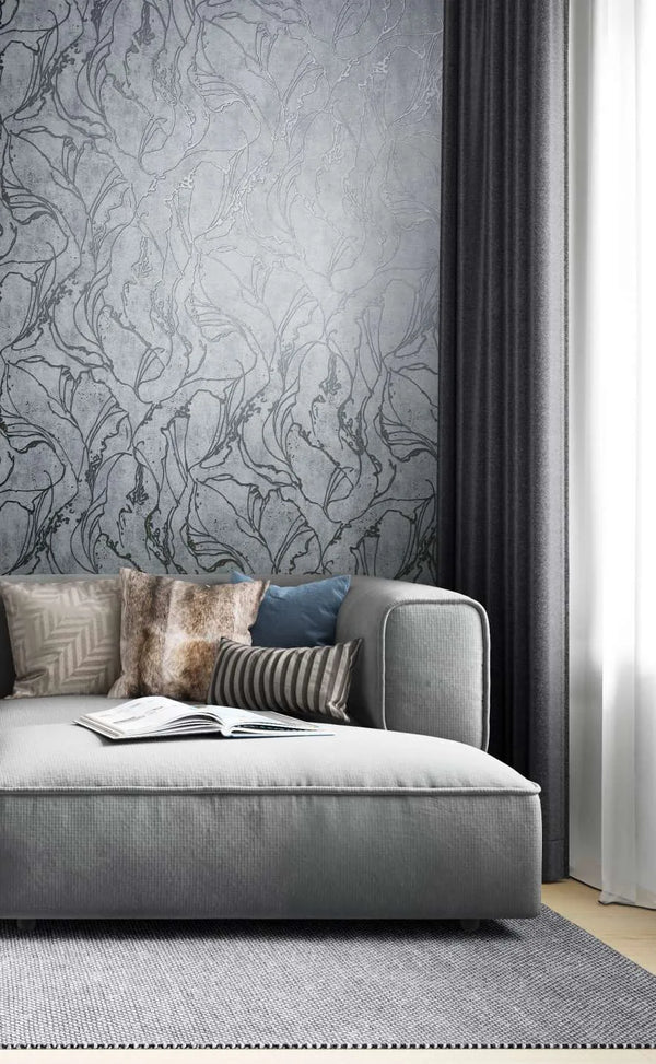 Marburg City Glow 34254 Gray wallpaper with a large flowing botanical line pattern behind a sofa with mixed pillows, next to dark curtains and a light rug; an open book rests on the seat.