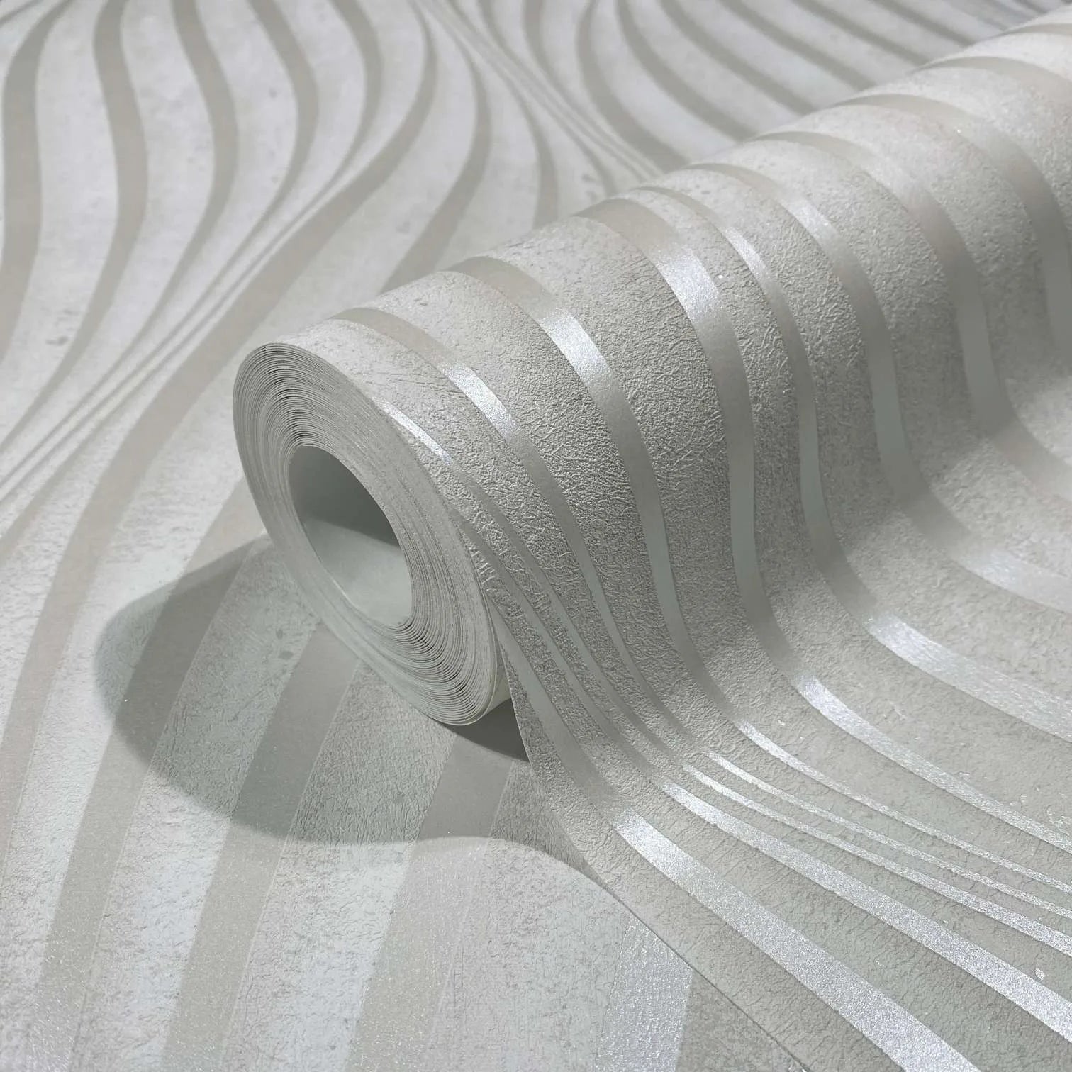 Marburg City Glow 34258 Close-up of a wallpaper roll on a matching sheet, showing light-toned wavy stripes with a subtle sheen and a softly textured background.