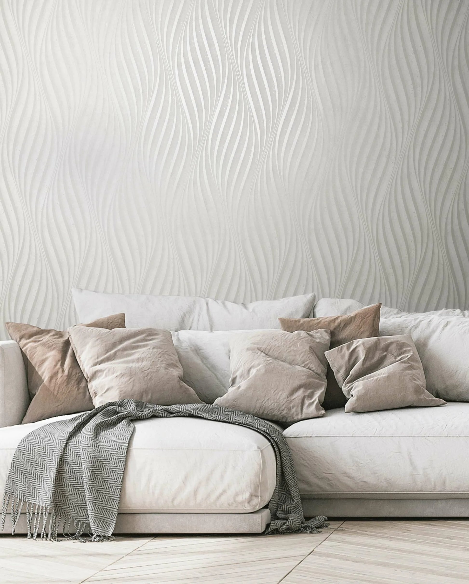 Marburg City Glow 34258 Living room scene showing light wavy-pattern wallpaper behind a soft sofa with neutral pillows and a draped throw blanket.