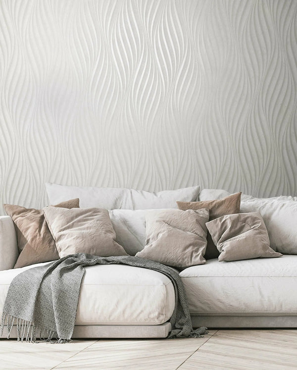 Marburg City Glow 34258 Living room scene showing light wavy-pattern wallpaper behind a soft sofa with neutral pillows and a draped throw blanket.