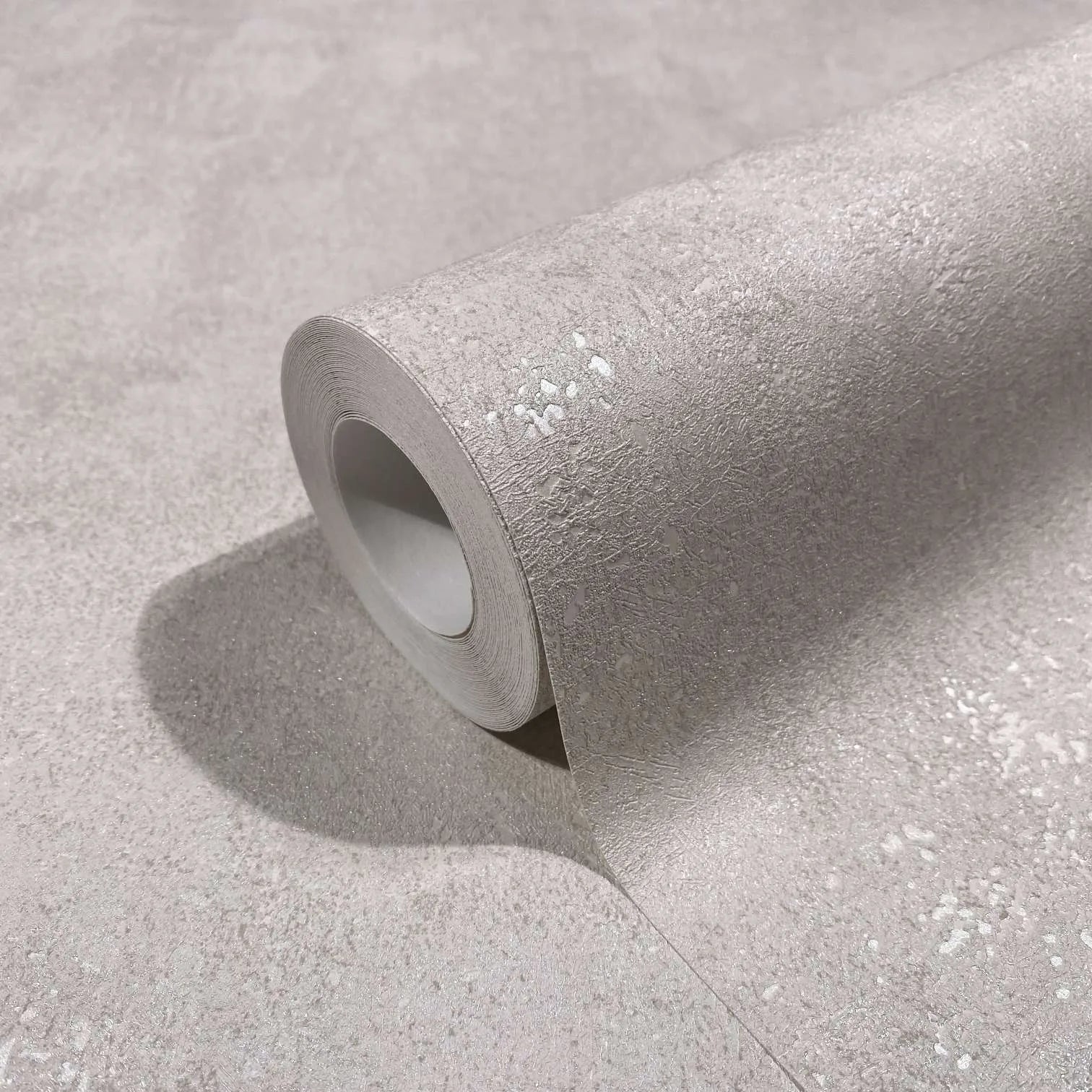 Marburg City Glow 34265 Close-up of a single roll of light beige textured wallpaper with fine irregular speckled highlights, partially unrolled on a matching surface.