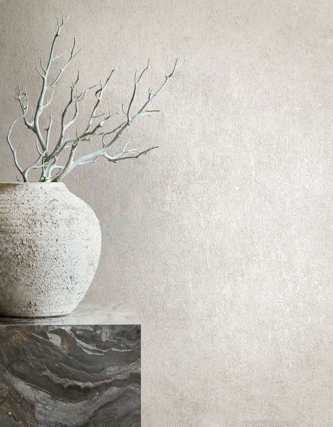 Marburg City Glow 34265 Beige textured wallpaper with a subtle plaster-like finish shown behind a rough stone vase holding bare branches on a dark marble pedestal.