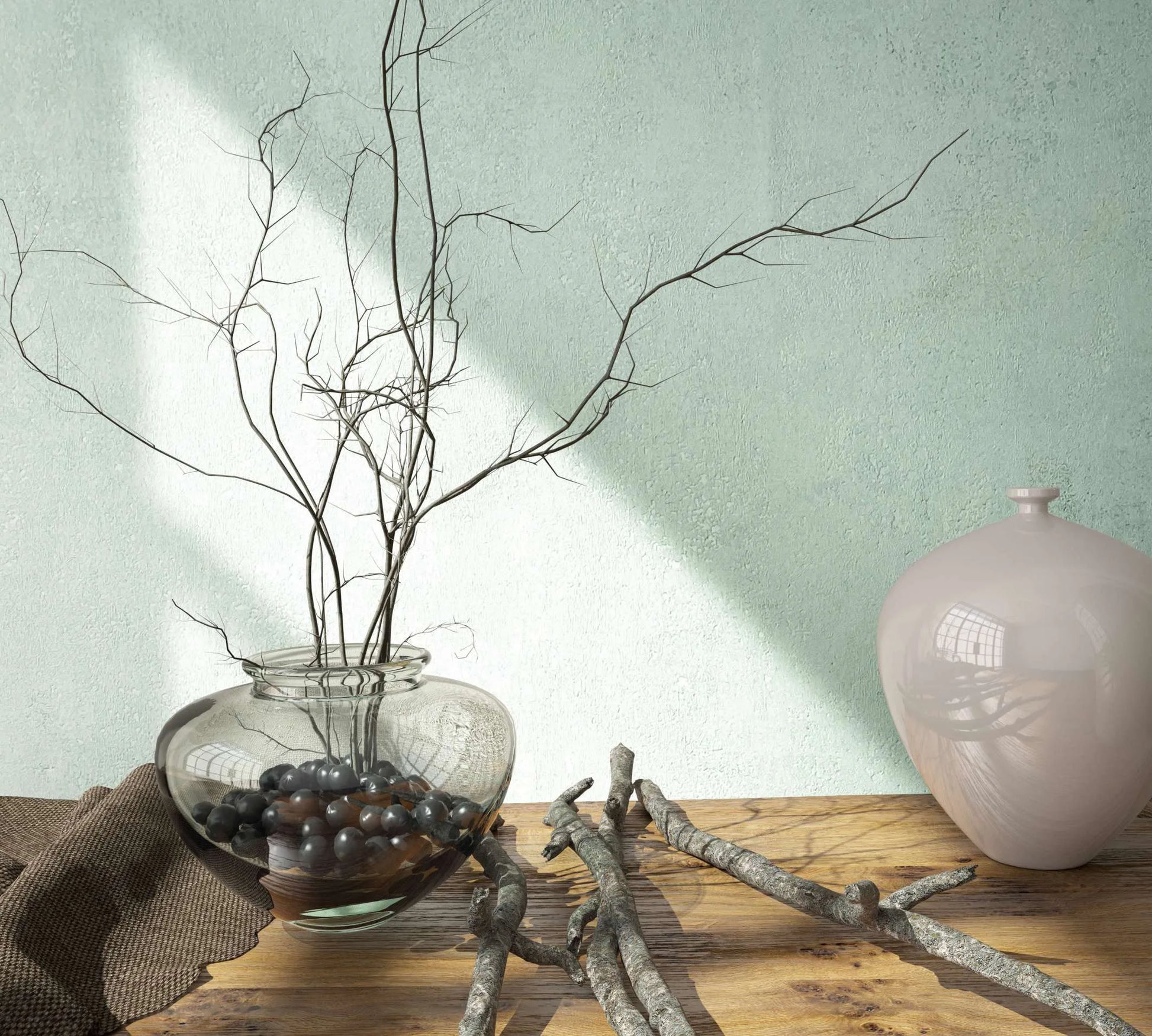 Marburg City Glow 34266 Light-toned, mottled textured wallpaper serves as a backdrop to a wooden tabletop with a glass vase holding branches and pebbles, a glossy ceramic vessel, scattered twigs, and a folded fabric, with sunlight casting diagonal shadows on the wall.