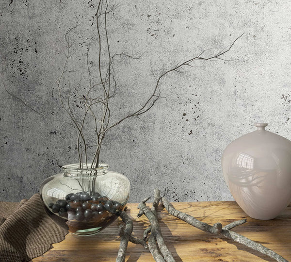 Marburg City Glow 34267 Gray concrete-look wallpaper behind a wood table styled with a clear glass vase of branches, a ceramic vase, scattered twigs, and a small cloth.