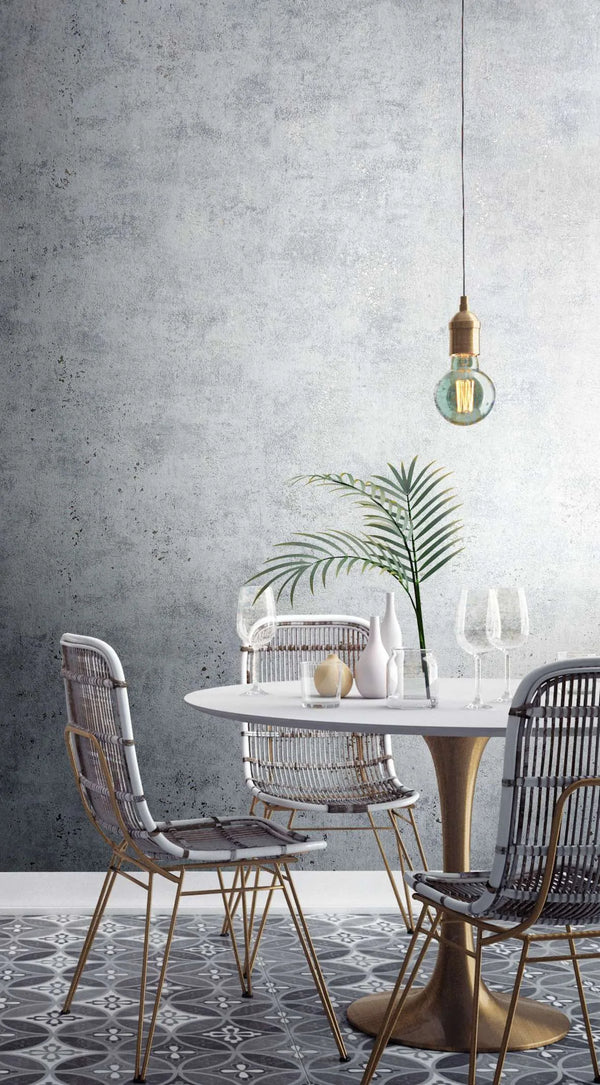 Marburg City Glow 34267 Gray concrete-look textured wallpaper behind a small dining area with a round table, woven chairs, a pendant bulb, glassware, a few vases, a green frond, and a patterned tile floor.
