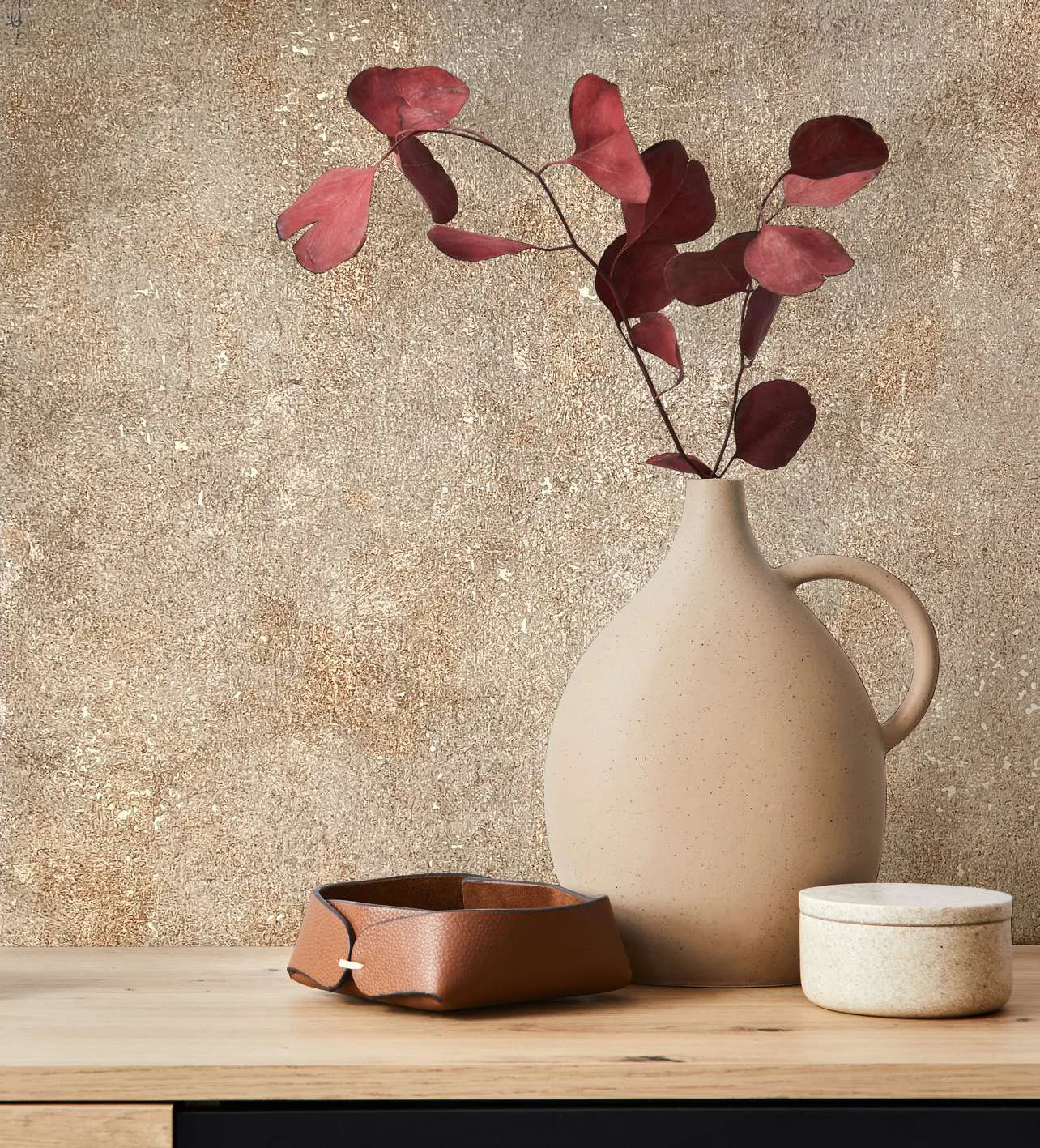 Marburg City Glow 34269 Mottled beige and brown wallpaper with a speckled texture behind a ceramic jug holding red leaves, a small leather tray, and a lidded pot on a wooden surface.