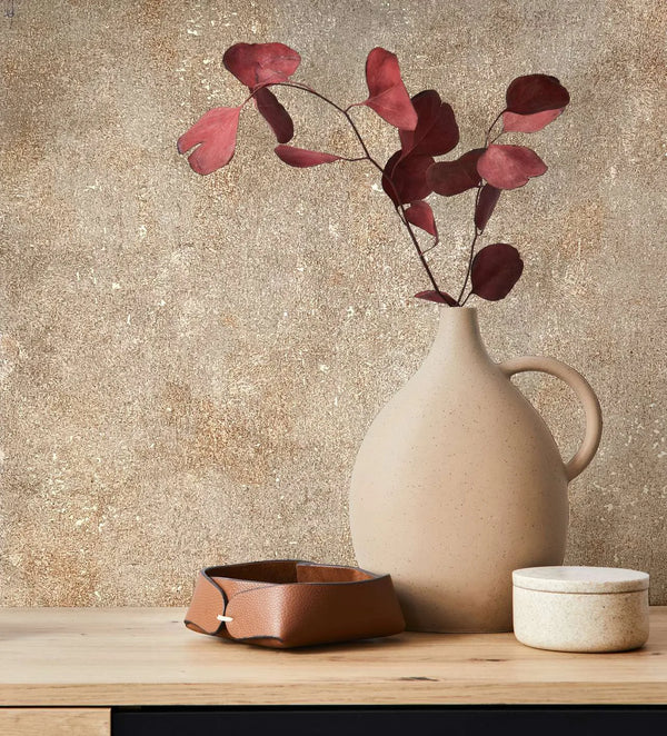 Marburg City Glow 34269 Mottled beige and brown wallpaper with a speckled texture behind a ceramic jug holding red leaves, a small leather tray, and a lidded pot on a wooden surface.