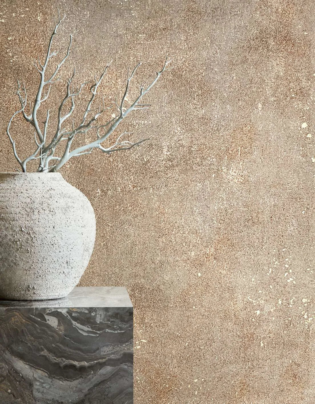 Marburg City Glow 34269 Speckled beige-brown textured wallpaper behind a rough stone vase with bare branches on a dark stone pedestal.