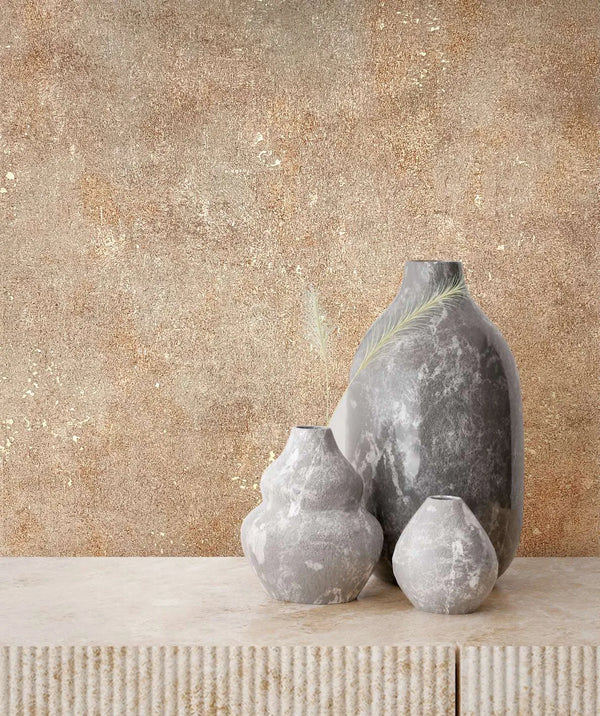 Marburg City Glow 34269 Textured beige and brown wallpaper with scattered gold flecks behind three gray stone vases on a ribbed stone console.