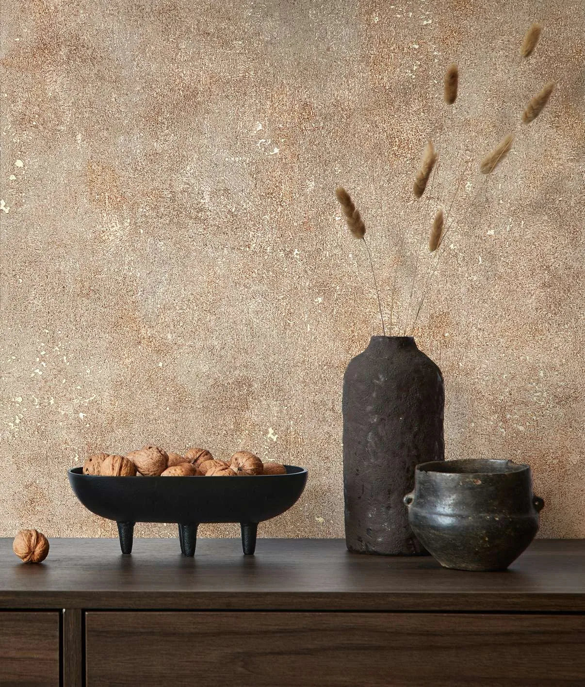 Marburg City Glow 34269 Warm beige and brown mottled wallpaper with subtle metallic flecks behind a wood sideboard holding a black tripod bowl of walnuts, a tall dark textured vase with dried stems, and a small ceramic pot.