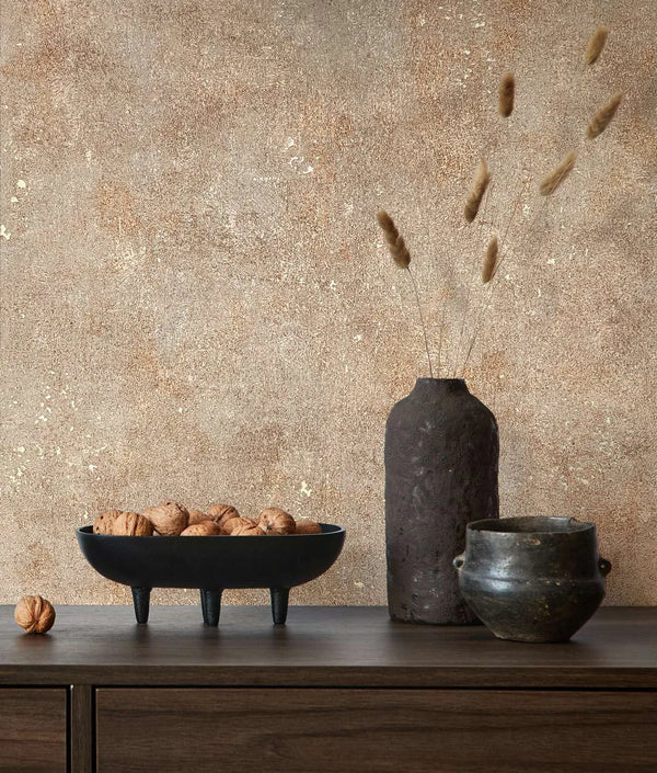 Marburg City Glow 34269 Warm beige and brown mottled wallpaper with subtle metallic flecks behind a wood sideboard holding a black tripod bowl of walnuts, a tall dark textured vase with dried stems, and a small ceramic pot.