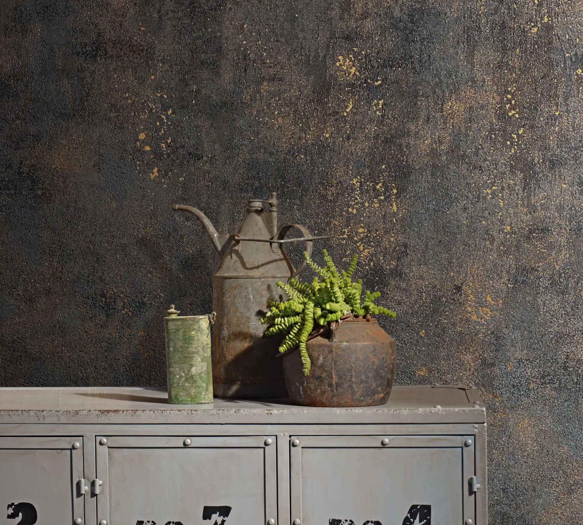Marburg City Glow 34270 Textured black wallpaper with copper flecks behind an industrial locker topped with a metal watering can, a small canister, and a potted fern.