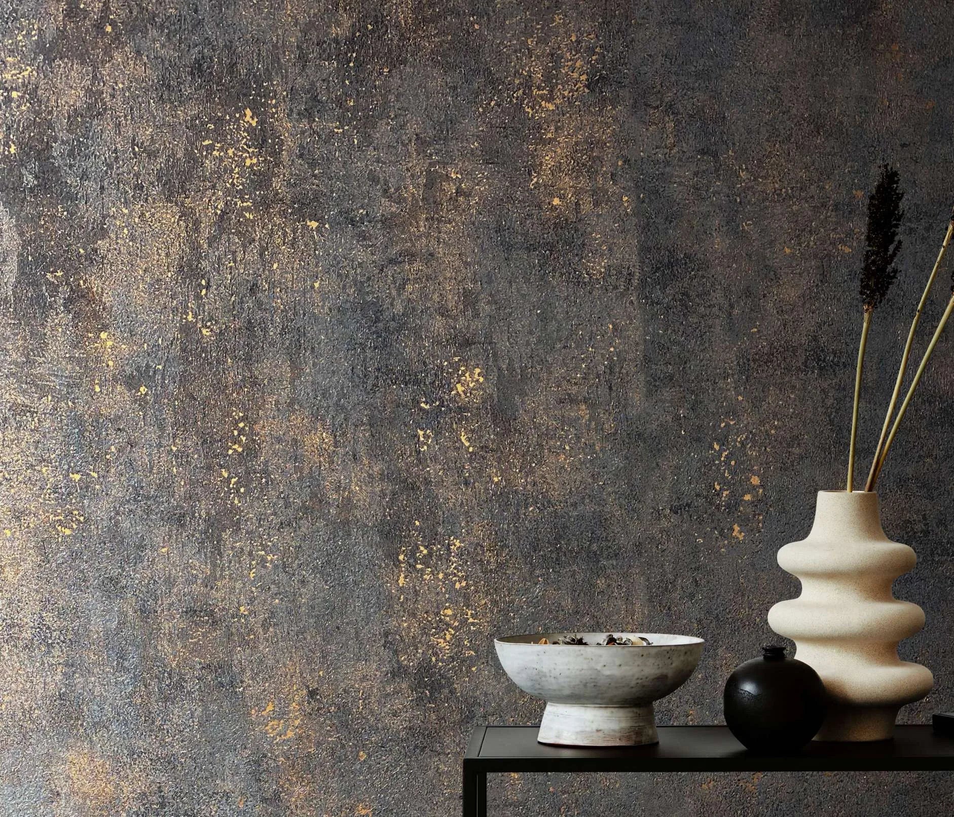 Marburg City Glow 34270 A dark charcoal textured wallpaper with scattered warm metallic flecks forms the backdrop to a black console holding a wavy cream vase with dried stems, a shallow gray bowl, and a small round black vase.
