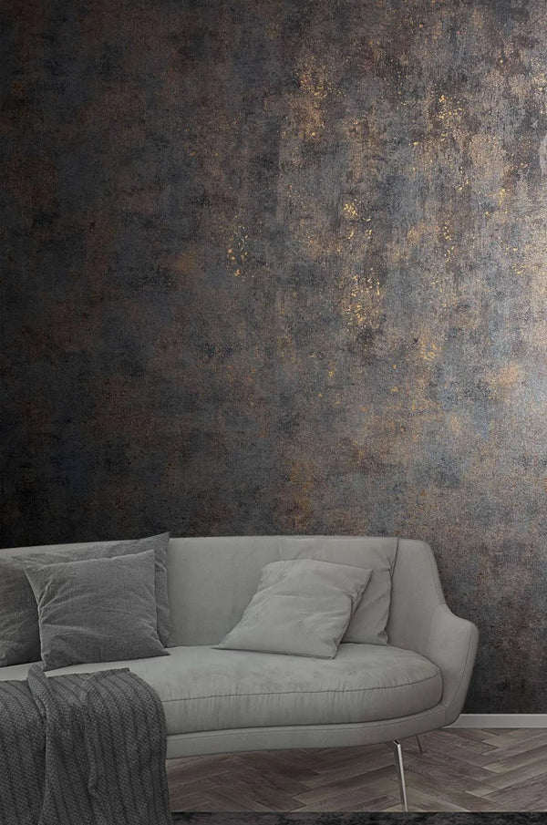 Marburg City Glow 34270 Room scene showing a dark charcoal wallpaper with copper-flecked, distressed texture behind a light gray curved sofa with pillows and a knit throw; baseboard and herringbone wood floor visible.