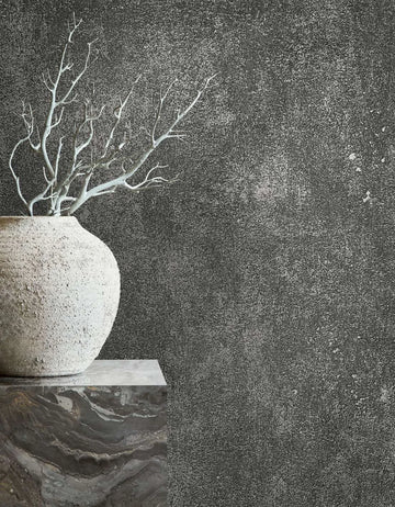 Marburg City Glow 34271 Dark charcoal textured wallpaper with a fine speckled, distressed finish. A rough stone vase holding bare branches sits on a marble pedestal at the left.