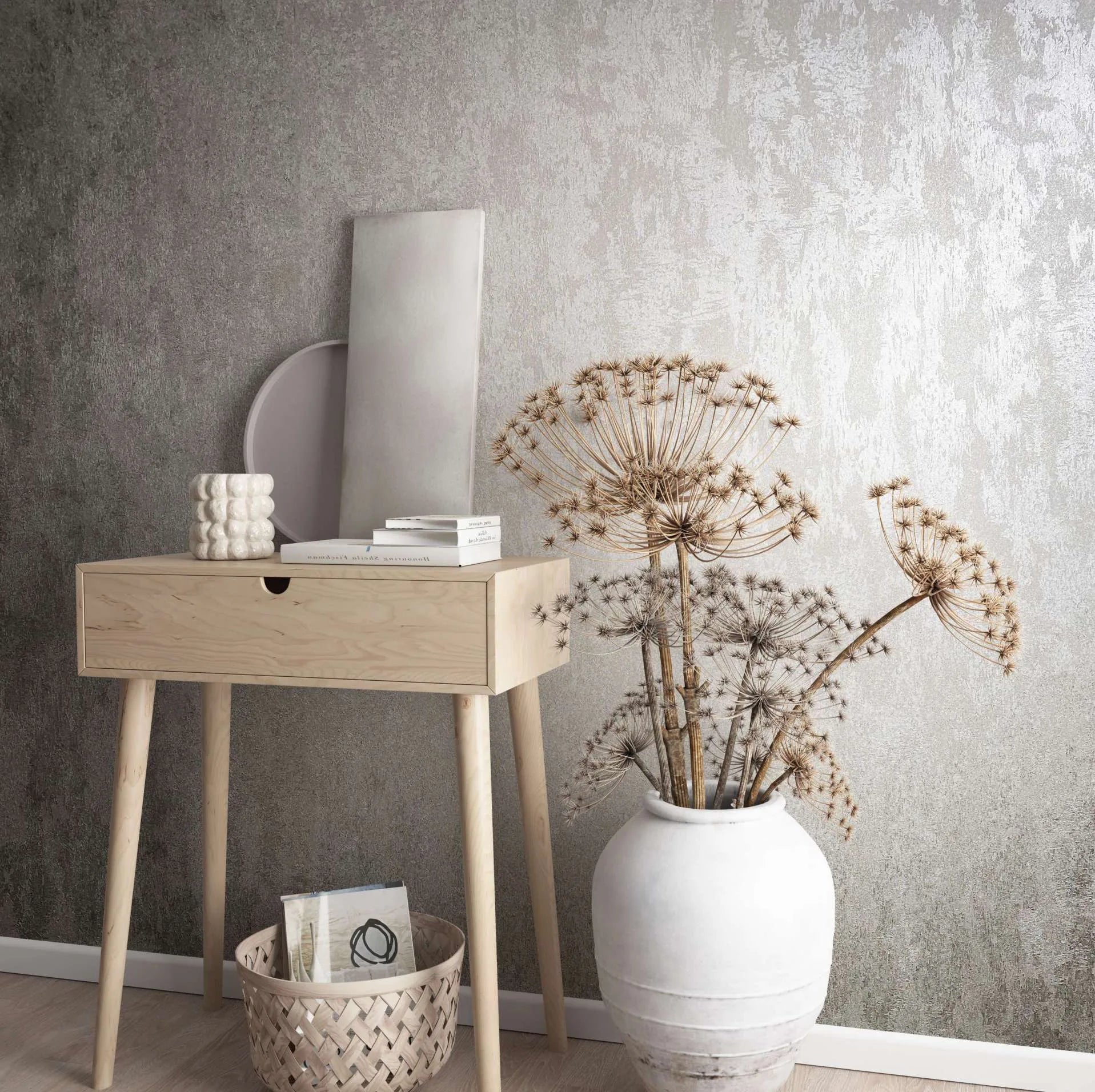 Marburg City Glow 34274 Abstract tonal gray wallpaper with a mottled, painterly texture shown behind a light wood console table with books and a sculptural candle, a leaning round tray and panel, a woven basket on the floor, and a large white ceramic vase holding dried seed heads.