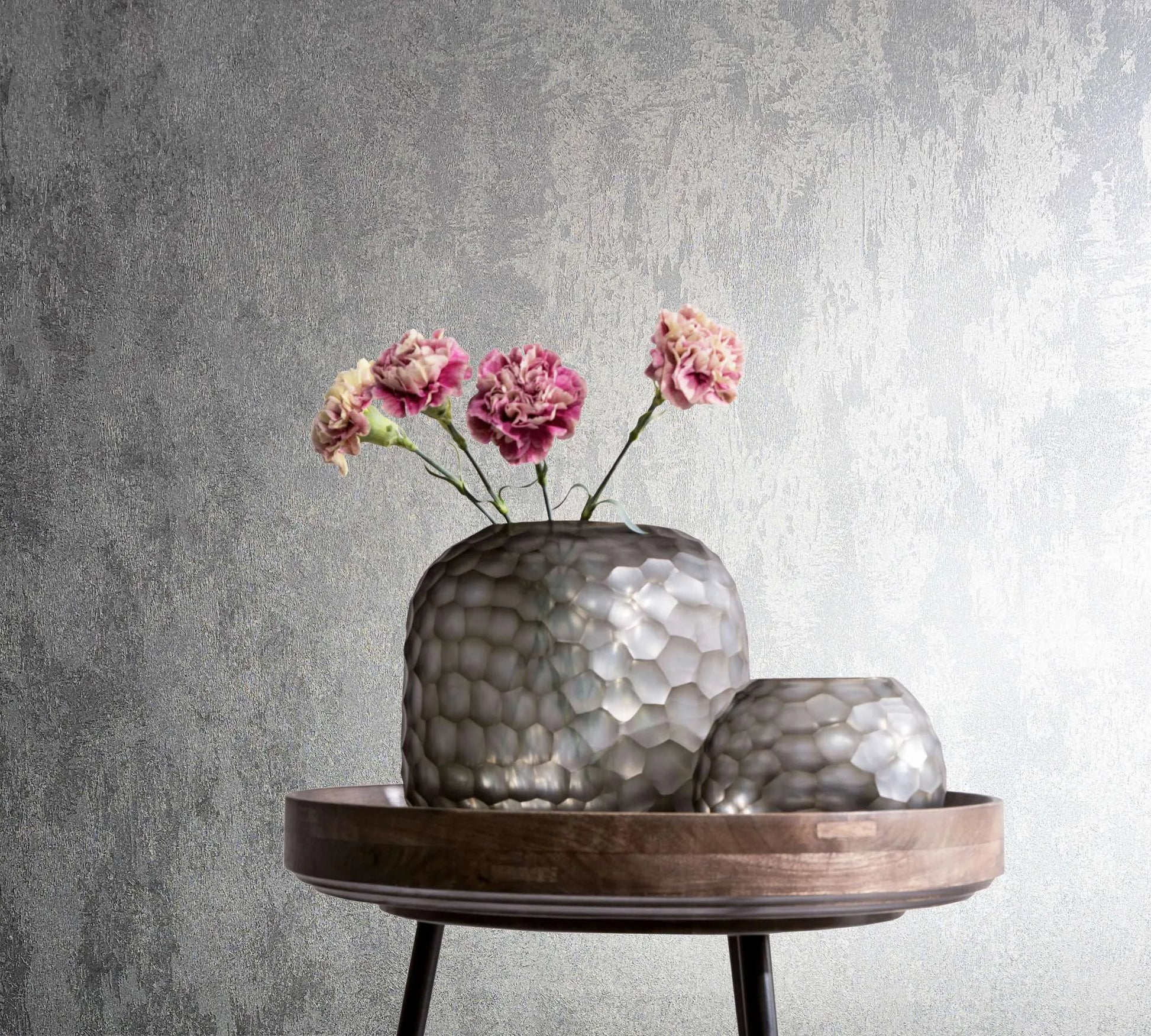 Marburg City Glow 34275 Textured silver wallpaper with an abstract brushed pattern, shown behind a round wooden tray table holding two faceted vases and pink flowers.