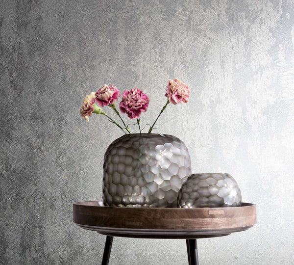 Marburg City Glow 34275 Textured silver wallpaper with an abstract brushed pattern, shown behind a round wooden tray table holding two faceted vases and pink flowers.
