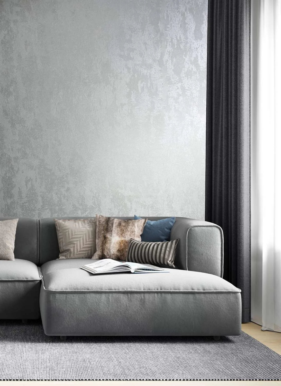Marburg City Glow 34275 Silver textured wallpaper with a subtle sheen behind a gray sectional sofa with mixed cushions, dark curtains, a sheer window panel, and an open book on the seat.