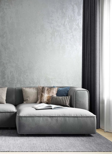 Marburg City Glow 34275 Silver textured wallpaper with a subtle sheen behind a gray sectional sofa with mixed cushions, dark curtains, a sheer window panel, and an open book on the seat.