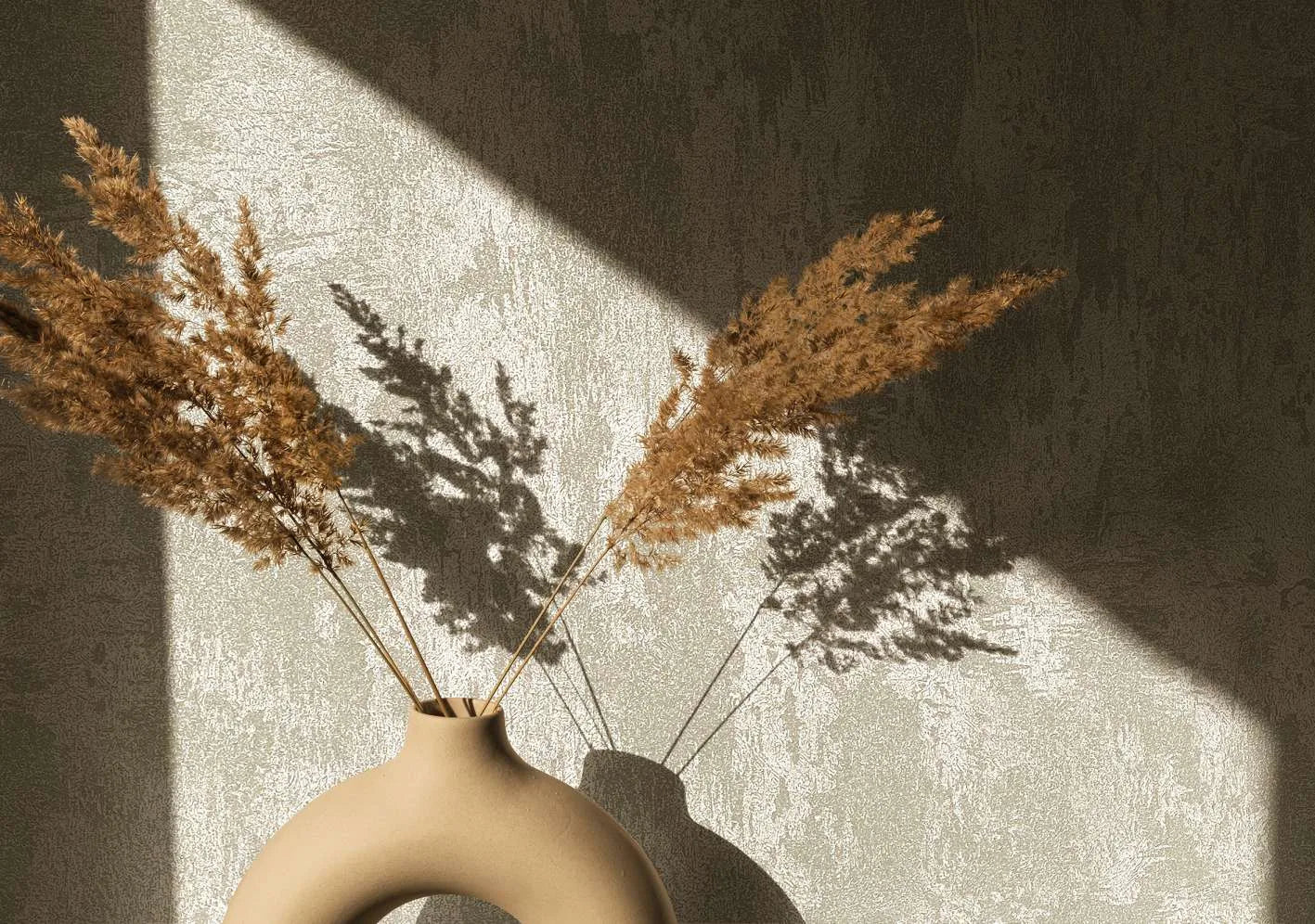 Marburg City Glow 34276 Close-up of textured light gray wallpaper with an irregular plaster-style pattern, lit by a diagonal beam of sunlight. A matte beige sculptural vase holds dried grasses, casting sharp shadows on the wall.