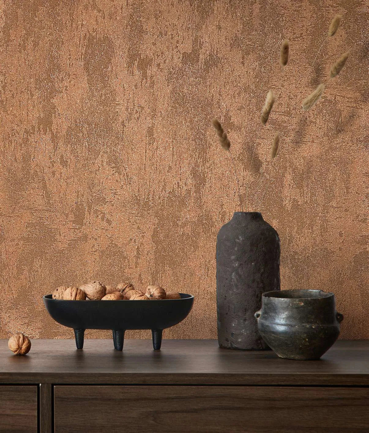 Marburg City Glow 34277 Textured copper wallpaper with a stucco-like pattern behind a wood sideboard styled with a black three-legged bowl of walnuts, a tall dark vase with dried stems, and a small pot.