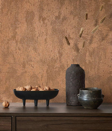 Marburg City Glow 34277 Textured copper wallpaper with a stucco-like pattern behind a wood sideboard styled with a black three-legged bowl of walnuts, a tall dark vase with dried stems, and a small pot.