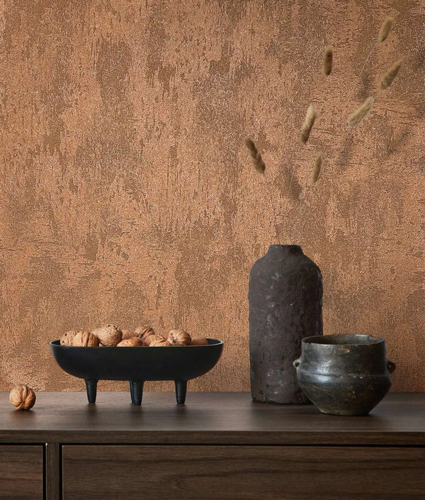 Marburg City Glow 34277 Textured copper wallpaper with a stucco-like pattern behind a wood sideboard styled with a black three-legged bowl of walnuts, a tall dark vase with dried stems, and a small pot.