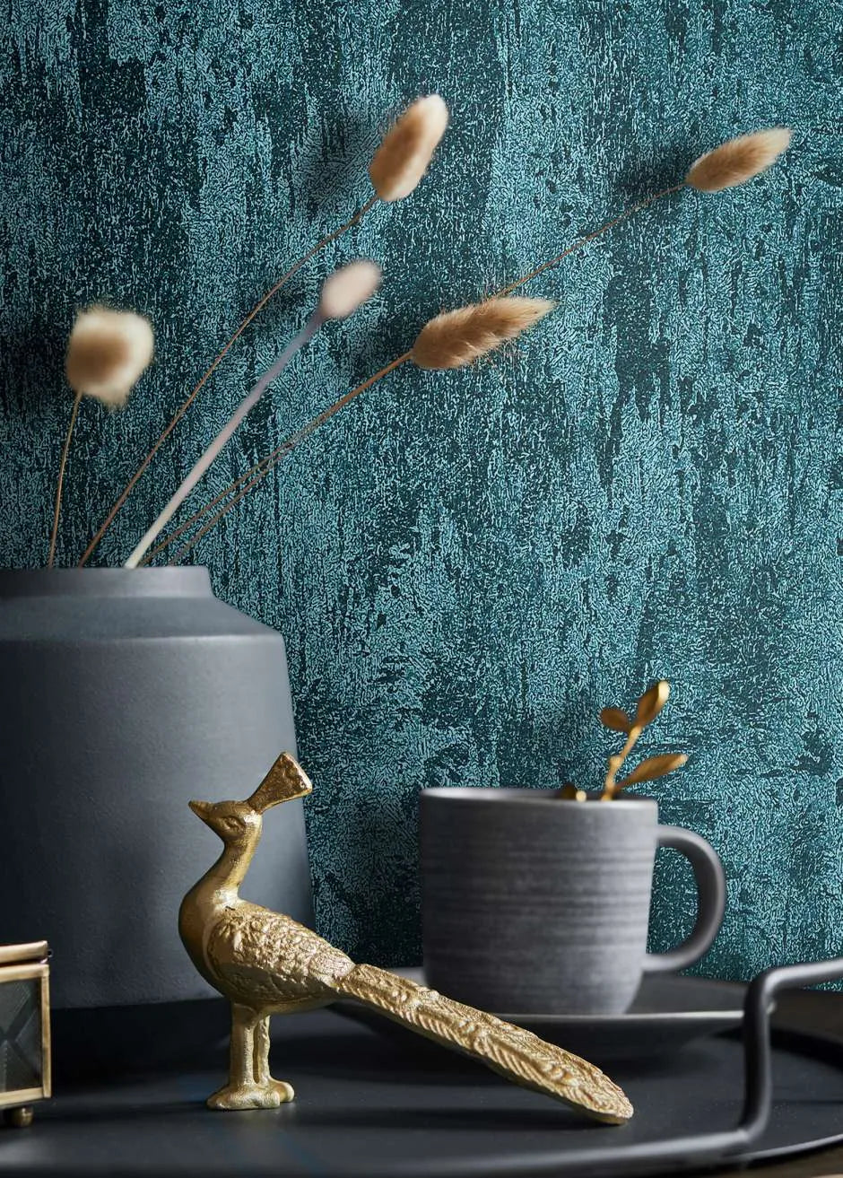 Marburg City Glow 34278 Close-up of blue-green textured wallpaper behind a matte gray vase with dried stems, a gray mug on a saucer, and a small gold bird figurine on a dark surface.