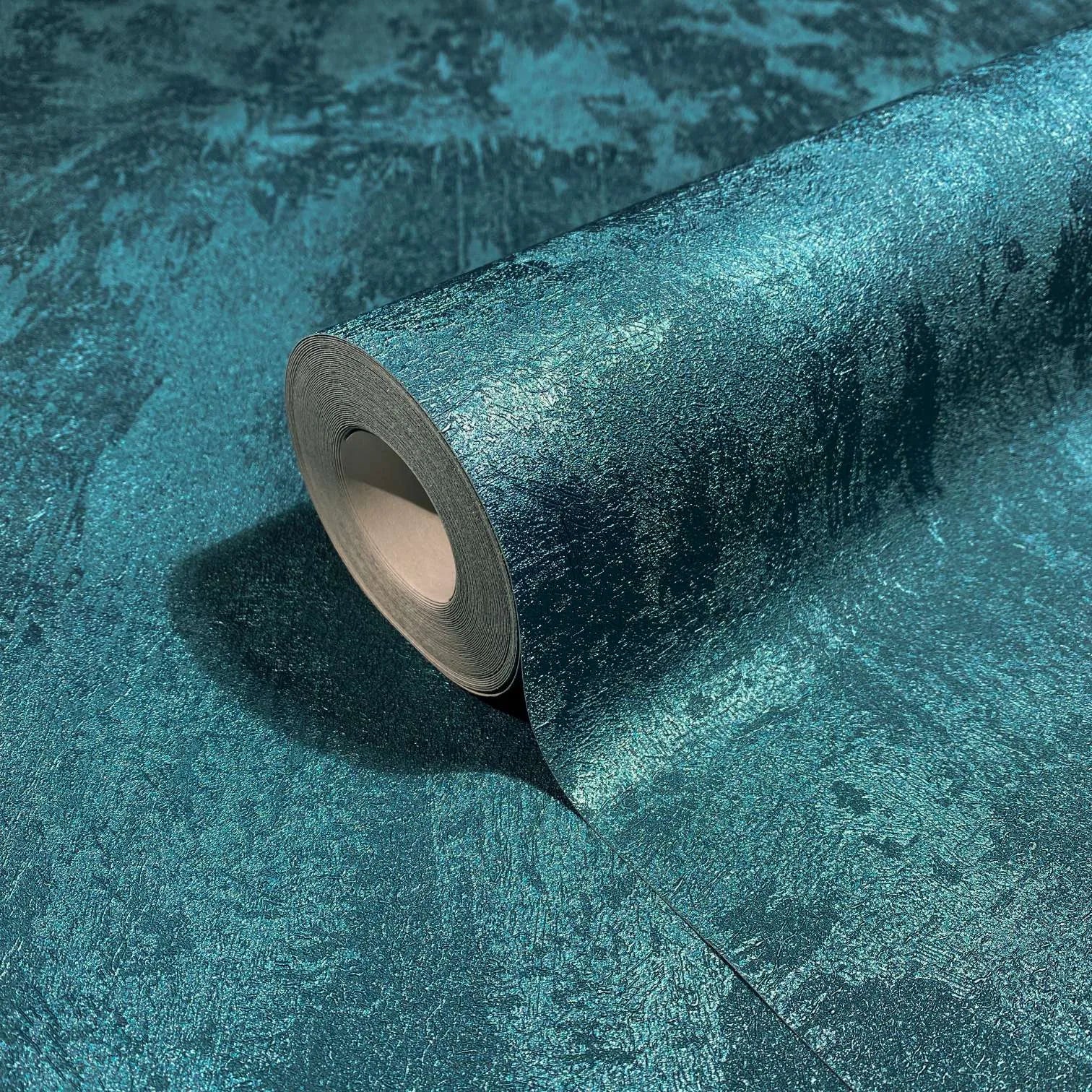 Marburg City Glow 34278 Close-up of a teal blue-green textured wallpaper with a roll resting on the same pattern, showing a mottled, subtly shimmery surface.