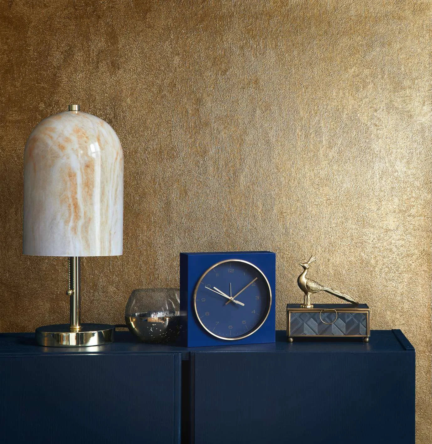 Marburg City Glow 34280 Metallic gold textured wallpaper behind a navy cabinet styled with a dome table lamp, a blue square clock, a small glass votive, and a gold bird figurine on a decorative box.