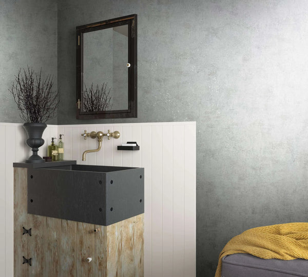 Marburg City Glow 34281 Bathroom scene showing gray mottled wallpaper above white vertical paneling, a vanity with a dark basin, wall-mounted mirror and faucet, small wall shelf with soap, bottles, a decorative urn with branches, and a gray ottoman with a yellow throw.